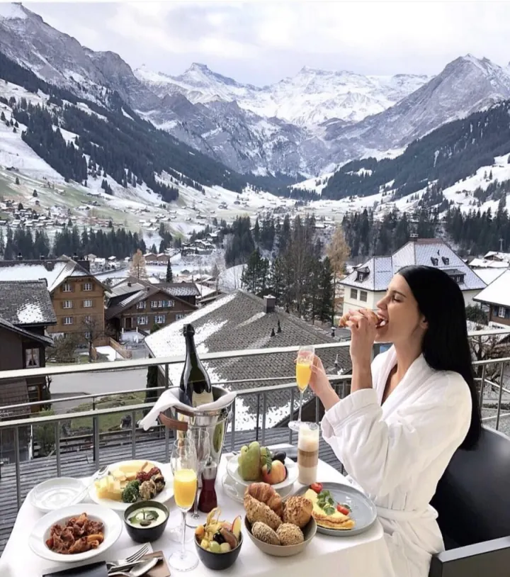 Shani in a white robe at breakfast on a balcony facing snowy Alpine peaks