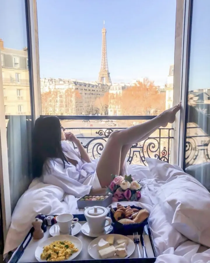 Shani in a white robe having breakfast in bed by an open window facing the Eiffel Tower
