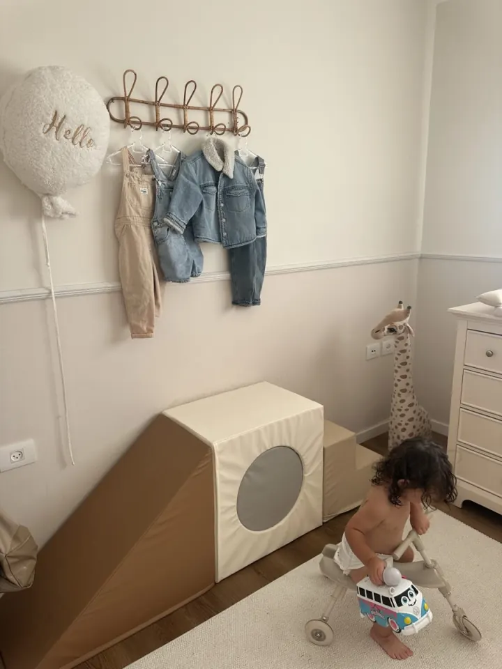 Toddler playing in a nursery corner with a soft slide and denim clothes on rattan hooks