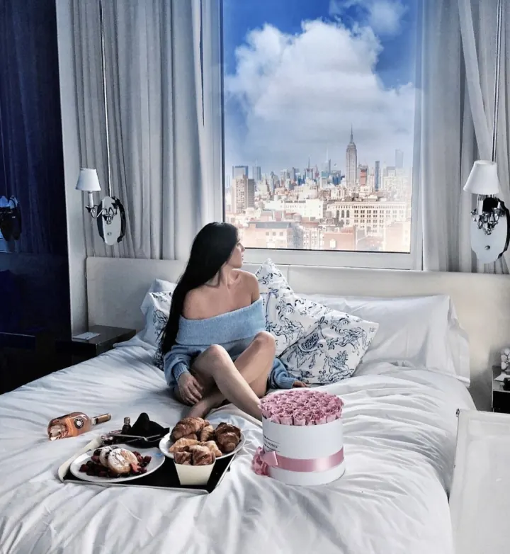 Shani on a hotel bed with breakfast and pink roses, the Manhattan skyline in the window