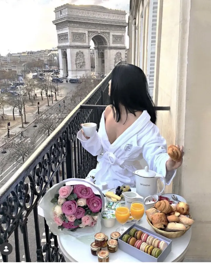 Shani in a robe having breakfast with flowers and macarons on a Paris balcony facing the Arc de Triomphe